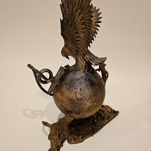 May include: A gold-colored eagle sculpture with its wings spread, perched atop a globe with a textured surface. The globe is surrounded by two anchors and a rope, and the sculpture is mounted on a base shaped like a continent.