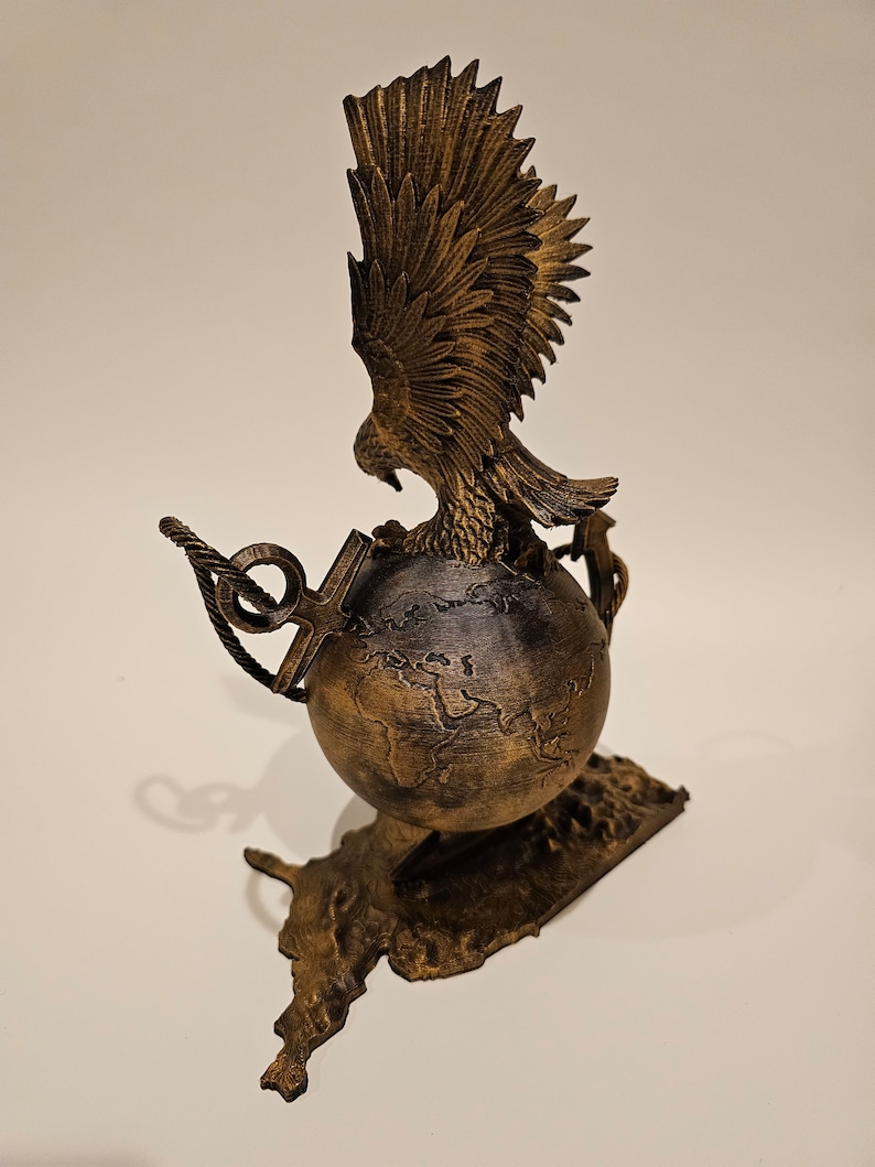 May include: A gold-colored eagle sculpture with its wings spread, perched atop a globe with a textured surface. The globe is surrounded by two anchors and a rope, and the sculpture is mounted on a base shaped like a continent.