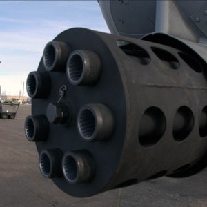 May include: Close-up of a gray aircraft cannon with multiple barrels. The cannon is mounted on a gray aircraft.