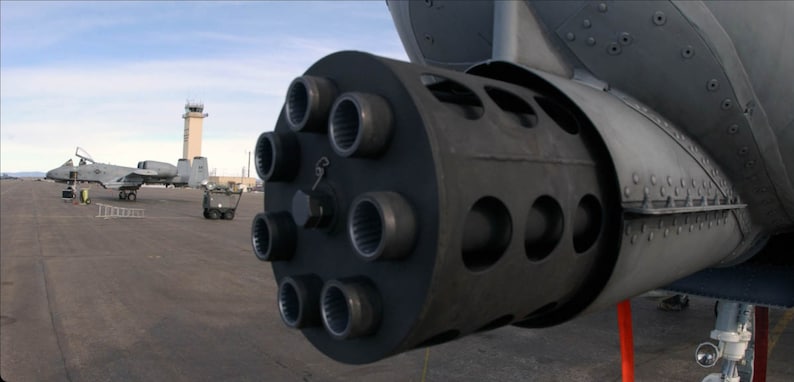 May include: Close-up of a gray aircraft cannon with multiple barrels. The cannon is mounted on a gray aircraft.