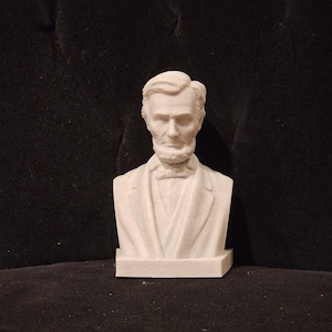 May include: White bust sculpture of Abraham Lincoln, a historical figure, with a serious expression. The bust is on a white base.