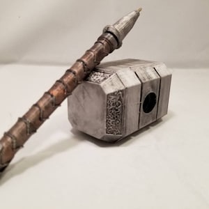 Op de afbeelding: Een zilveren en bruine pen in de vorm van Thor's hamer, Mjolnir. De pen ligt op een wit oppervlak.