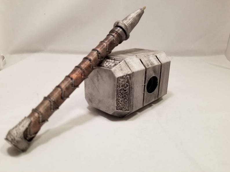 Op de afbeelding: Een zilveren en bruine pen in de vorm van Thor's hamer, Mjolnir. De pen ligt op een wit oppervlak.