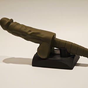 May include: A sculpture of a stylized phallus in a muted olive green color, mounted on a black base. The sculpture has a textured surface and is positioned at an angle, casting a shadow.