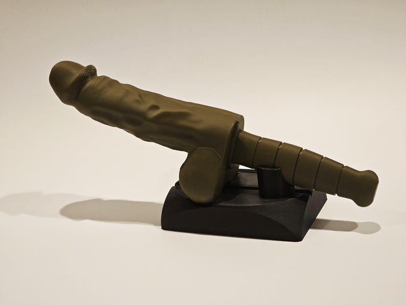 May include: A sculpture of a stylized phallus in a muted olive green color, mounted on a black base. The sculpture has a textured surface and is positioned at an angle, casting a shadow.