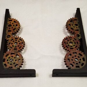 Puede incluir: Dos soportes de estanter&iacute;a de metal negro con un dise&ntilde;o decorativo de cuatro engranajes apilados. Los engranajes est&aacute;n pintados en tonos de naranja, amarillo y marr&oacute;n.
