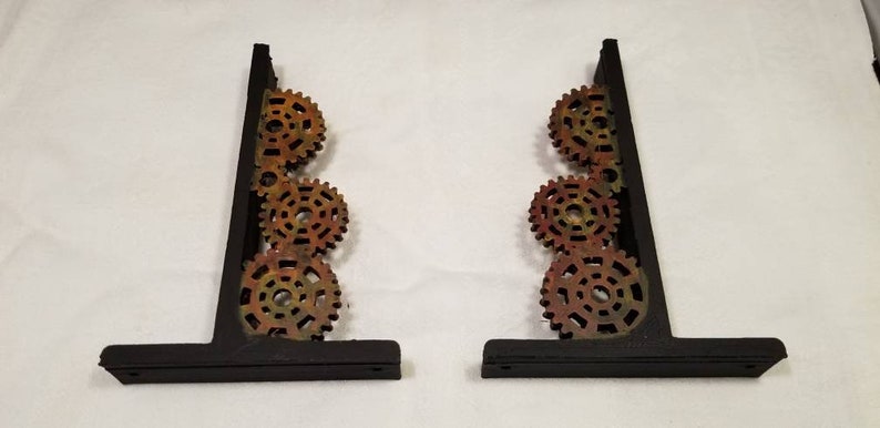 Puede incluir: Dos soportes de estanter&iacute;a de metal negro con un dise&ntilde;o decorativo de cuatro engranajes apilados. Los engranajes est&aacute;n pintados en tonos de naranja, amarillo y marr&oacute;n.