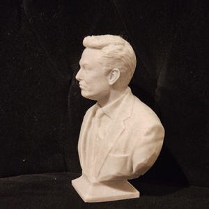 May include: A white bust sculpture of a person with short, styled hair. The person is wearing a suit jacket and a tie. The bust is on a white base.