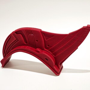 Op de afbeelding: Een rood plastic 3D-geprint onderdeel met een gebogen vorm en ingewikkelde geometrische ontwerpen. Het onderdeel heeft een gat aan de zijkant en een verhoogd gedeelte aan de bovenkant.