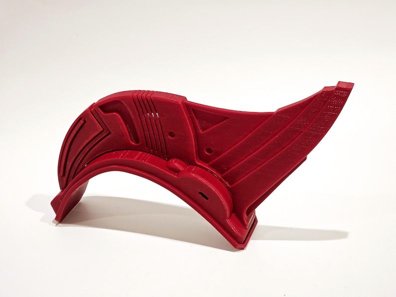 Op de afbeelding: Een rood plastic 3D-geprint onderdeel met een gebogen vorm en ingewikkelde geometrische ontwerpen. Het onderdeel heeft een gat aan de zijkant en een verhoogd gedeelte aan de bovenkant.