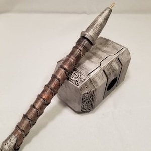 Op de afbeelding: Een zilveren en bruine pen in de vorm van Thor's hamer, Mjolnir. De pen ligt op een zilveren en grijze, hamer-vormige pennenhouder.