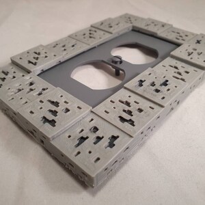 Peut inclure: Une plaque de prise de courant grise imprim&eacute;e en 3D avec un design textur&eacute;. La plaque est rectangulaire et compos&eacute;e de petits carr&eacute;s avec des trous.