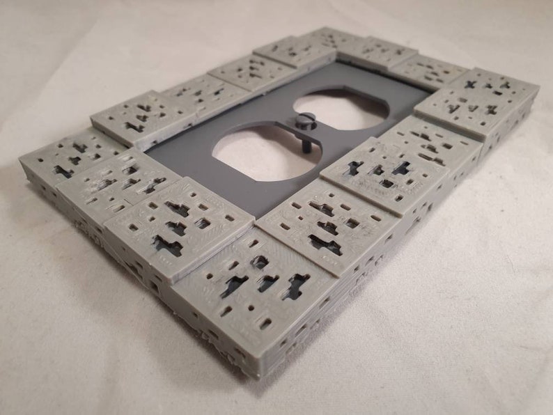 Peut inclure: Une plaque de prise de courant grise imprim&eacute;e en 3D avec un design textur&eacute;. La plaque est rectangulaire et compos&eacute;e de petits carr&eacute;s avec des trous.