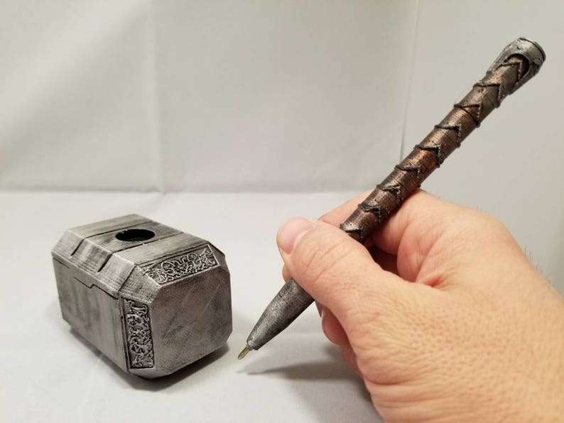 Op de afbeelding: Een zilveren metalen pennenhouder in de vorm van Thor's hamer, Mjolnir, met ingewikkelde details. Naast de houder ligt een bruine en zilveren pen met een gevlochten ontwerp.