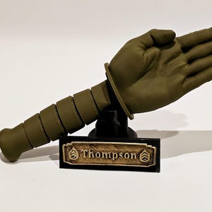 May include: A green 3D printed model of a hand giving a salute, mounted on a black base with a gold nameplate that reads "Thompson".