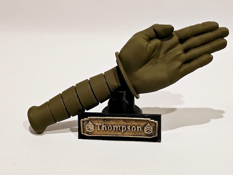 May include: A green 3D printed model of a hand giving a salute, mounted on a black base with a gold nameplate that reads "Thompson".