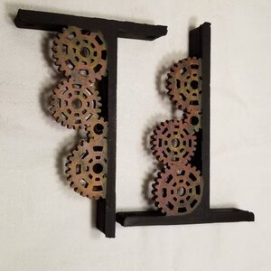 Puede incluir: Dos sujetalibros de metal negro con un dise&ntilde;o decorativo de engranajes entrelazados en tonos marrones, dorados y rojos.