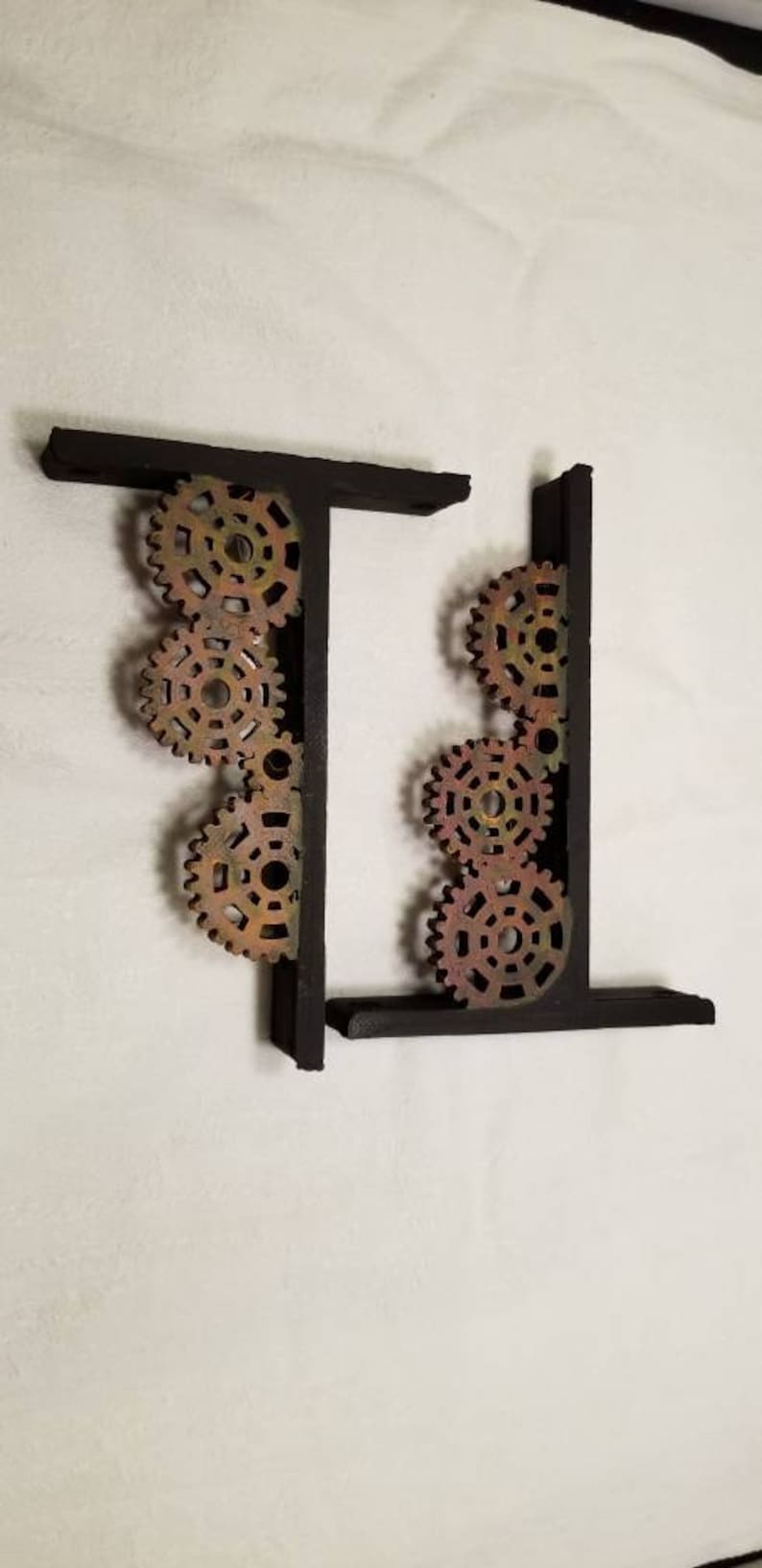 Puede incluir: Dos sujetalibros de metal negro con un dise&ntilde;o decorativo de engranajes entrelazados en tonos marrones, dorados y rojos.