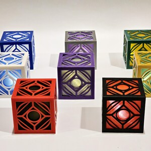 K&ouml;nnte beinhalten: Acht kleine, w&uuml;rfelf&ouml;rmige Boxen mit komplizierten geometrischen Mustern. Die Boxen sind in verschiedenen Farben, darunter Wei&szlig;, Rot, Schwarz, Lila, Grau, Blau, Gr&uuml;n und Gold.