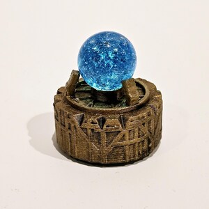 May include: A bright blue crackle glass sphere sits atop a circular, bronze-coloured base. The base features a textured, architectural design with vertical supports and a gear-like rim, holding the sphere securely in the centre of the structure.