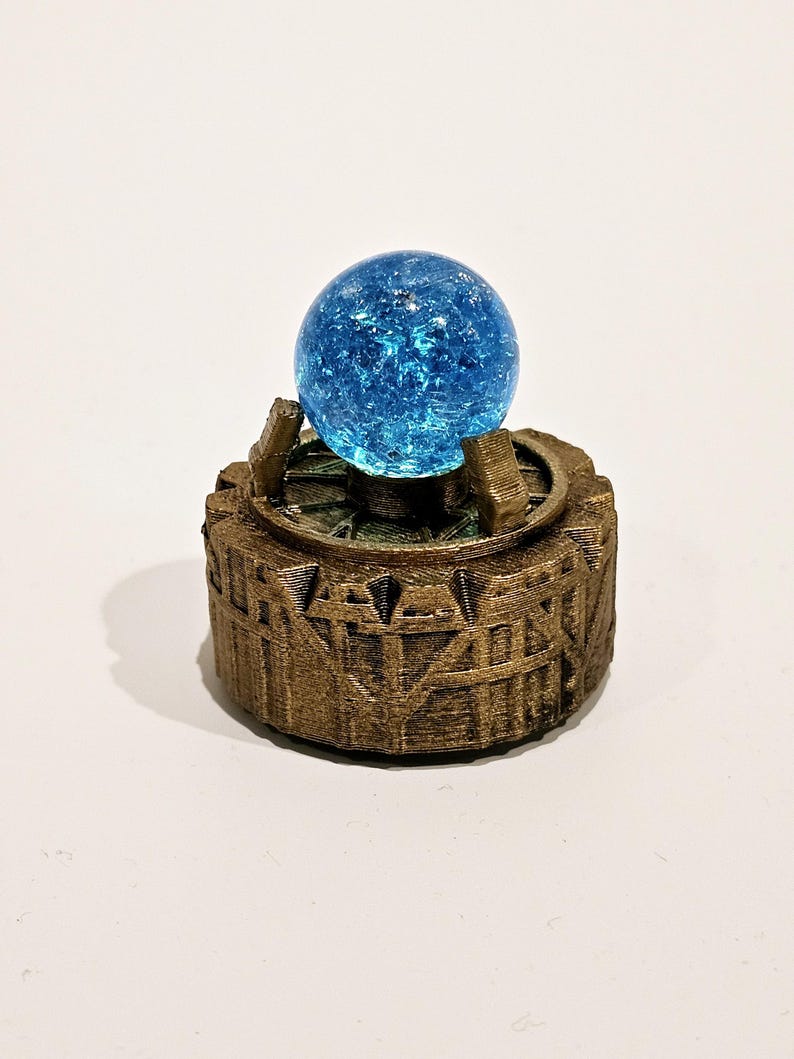 May include: A bright blue crackle glass sphere sits atop a circular, bronze-coloured base. The base features a textured, architectural design with vertical supports and a gear-like rim, holding the sphere securely in the centre of the structure.
