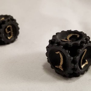 May include: Two black dice with gold numbers. The dice have a textured surface and are designed to look like gears.
