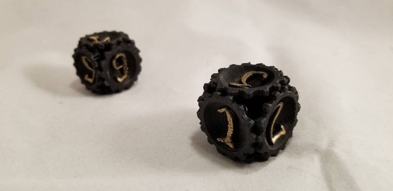 May include: Two black dice with gold numbers. The dice have a textured surface and are designed to look like gears.