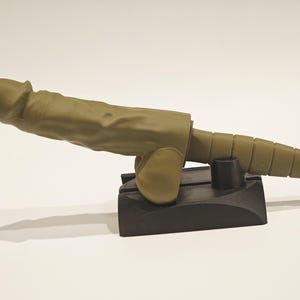 May include: A novelty item resembling a cannon, with a phallic design. The item is olive green and rests on a black base. The cannon barrel has a segmented design.