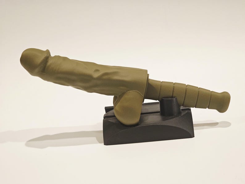 May include: A novelty item resembling a cannon, with a phallic design. The item is olive green and rests on a black base. The cannon barrel has a segmented design.