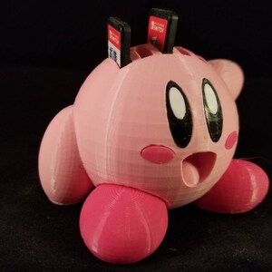 K&ouml;nnte beinhalten: Eine rosa 3D-gedruckte Kirby-Figur mit weit offenem Mund, die vier Nintendo Switch-Spielpatronen oben auf dem Kopf h&auml;lt.