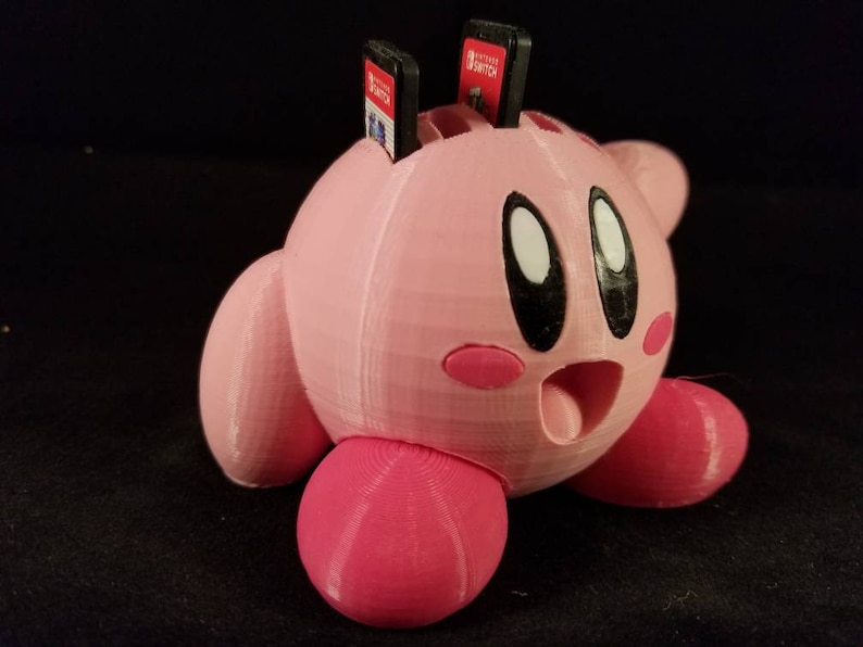 K&ouml;nnte beinhalten: Eine rosa 3D-gedruckte Kirby-Figur mit weit offenem Mund, die vier Nintendo Switch-Spielpatronen oben auf dem Kopf h&auml;lt.