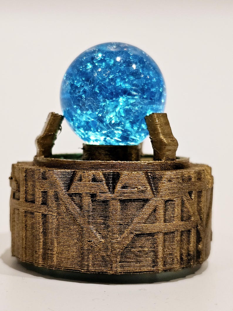May include: A bright blue, textured sphere rests atop a cylindrical, metallic bronze-coloured base. The base features a raised, geometric, and linear pattern around its circumference, with two small, upright supports holding the sphere in place.