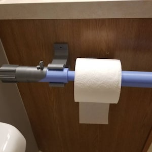 May include: A novelty toilet paper holder designed to resemble a lightsaber. The holder is gray and blue, with a roll of white toilet paper in place. The holder is mounted on a wooden surface.