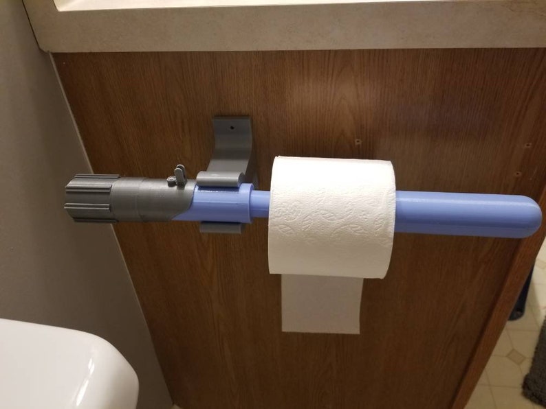 May include: A novelty toilet paper holder designed to resemble a lightsaber. The holder is gray and blue, with a roll of white toilet paper in place. The holder is mounted on a wooden surface.