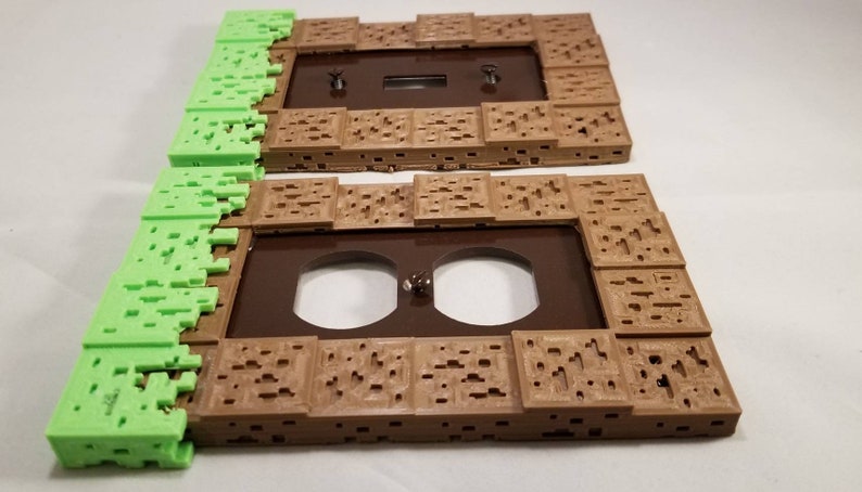 May include: Two 3D printed wall plates, one with a single light switch and the other with a double outlet. The plates are brown and have a textured design resembling a Minecraft block. The edges of the plates are green and resemble grass.