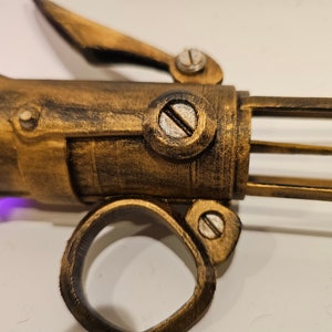 May include: A gold-colored, steampunk-style, 3D printed prop weapon with a purple light emanating from the barrel. The weapon has a ring handle and a lever on the top.