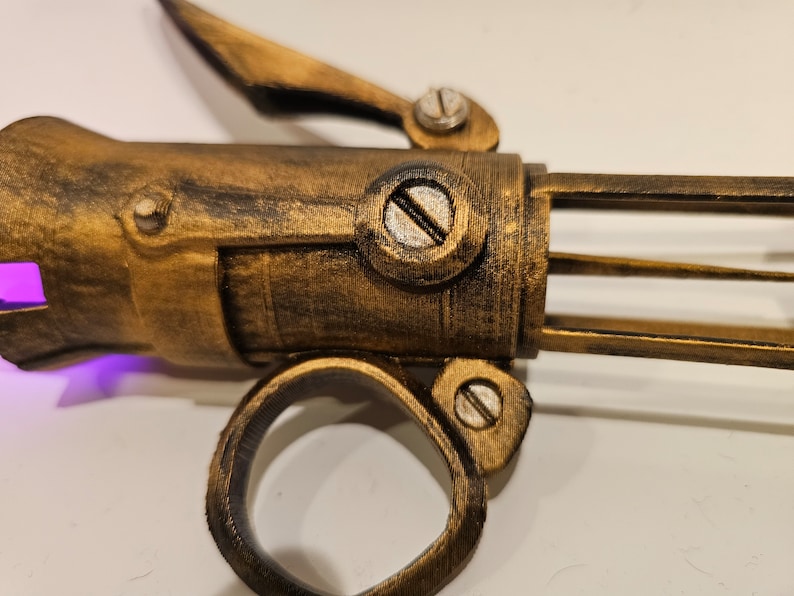 May include: A gold-colored, steampunk-style, 3D printed prop weapon with a purple light emanating from the barrel. The weapon has a ring handle and a lever on the top.