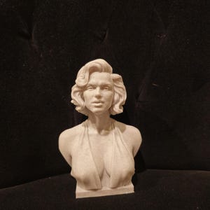 May include: A white bust sculpture of a woman with short, wavy hair. The sculpture is wearing a low-cut, sleeveless dress.