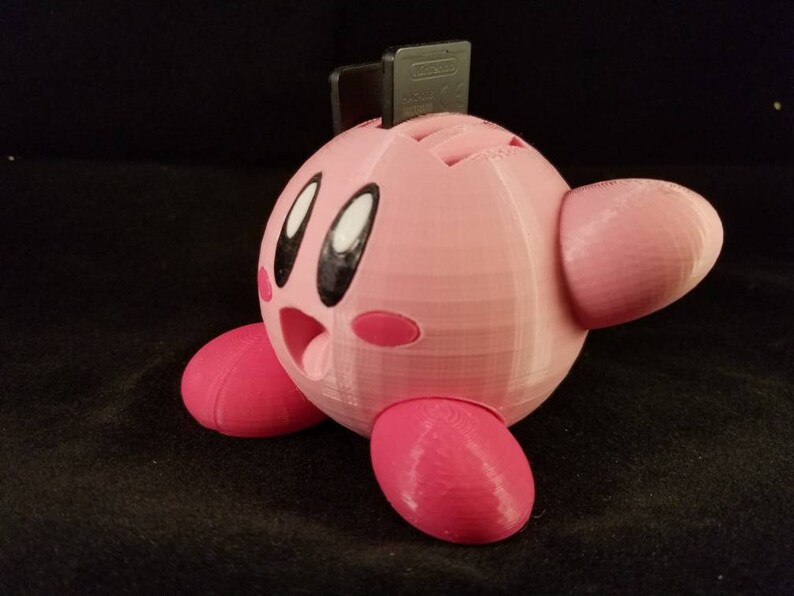 K&ouml;nnte beinhalten: Eine rosafarbene 3D-gedruckte Figur des Videospielcharakters Kirby. Kirby h&auml;lt eine schwarze Spielkassette in der Hand.
