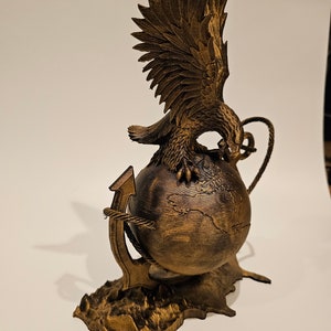 May include: A golden eagle sculpture with its wings spread, perched atop a globe with an anchor wrapped around it. The base is textured to resemble a rocky surface.