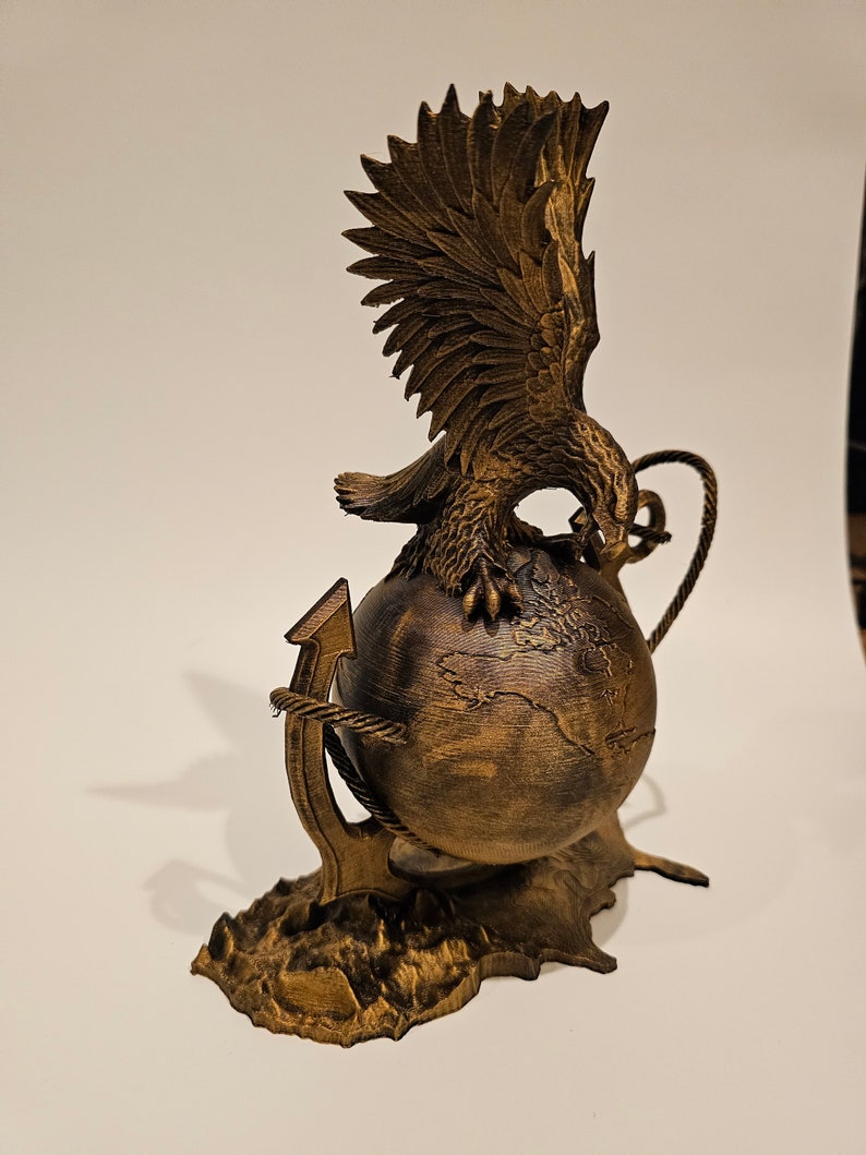 May include: A golden eagle sculpture with its wings spread, perched atop a globe with an anchor wrapped around it. The base is textured to resemble a rocky surface.