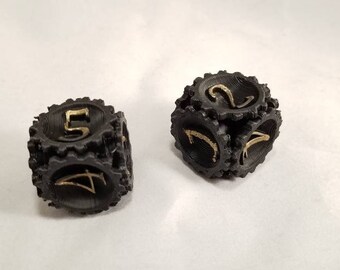 May include: Two black dice with gold numbers. The dice are in focus and have a textured surface. The dice are on a white background.