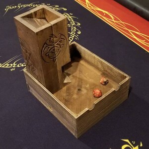 May include: Wooden dice tower with a dragon design. The tower has a slot for dice to fall into a wooden tray. Two red dice are in the tray.