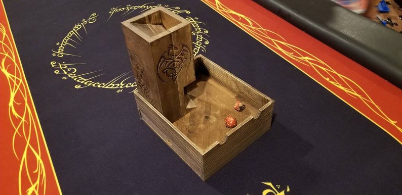May include: Wooden dice tower with a dragon design. The tower has a slot for dice to fall into a wooden tray. Two red dice are in the tray.