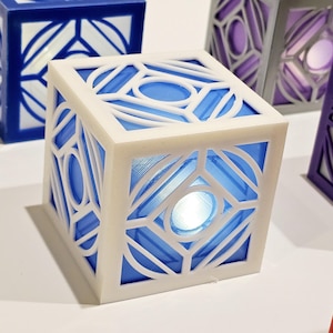 K&ouml;nnte beinhalten: Ein wei&szlig;er W&uuml;rfel mit einem blauen geometrischen Muster und einem wei&szlig;en Licht im Inneren. Der W&uuml;rfel ist aus Kunststoff und hat eine glatte Oberfl&auml;che.