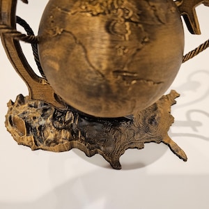 May include: A wooden globe with a rope handle and an anchor base. The globe is brown and has a textured surface. The base is shaped like a continent.