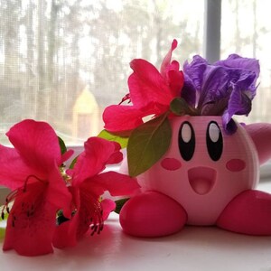 May include: A pink 3D printed planter shaped like the video game character Kirby, with a purple flower and pink flowers inside.