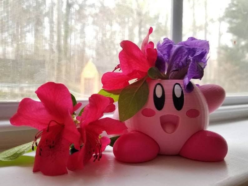 May include: A pink 3D printed planter shaped like the video game character Kirby, with a purple flower and pink flowers inside.