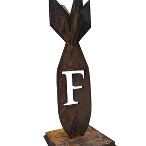 May include: A brown, stylized bomb-shaped sculpture with the letter "F" cut out. The sculpture has a textured surface and sits on a square base. The design features a fin-like structure at the top.