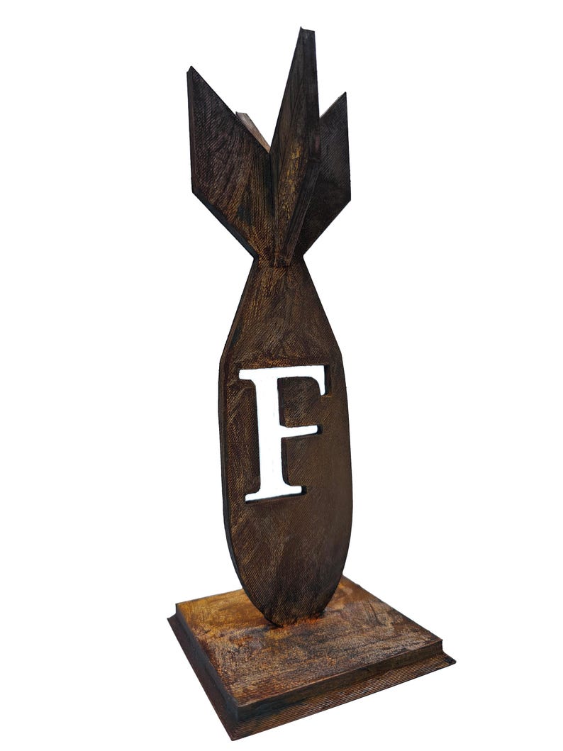 May include: A brown, stylized bomb-shaped sculpture with the letter "F" cut out. The sculpture has a textured surface and sits on a square base. The design features a fin-like structure at the top.